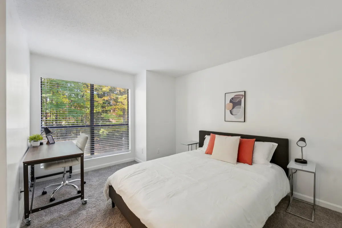 The image showcases a bright, inviting bedroom featuring a comfortable bed adorned with white bedding and vibrant red accent pillows. A desk with a chair sits near a large window, which bathes the room in natural light and offers peaceful views of lush greenery outside. Tasteful artwork on the wall adds a touch of personality, creating a harmonious blend of relaxation and productivity.