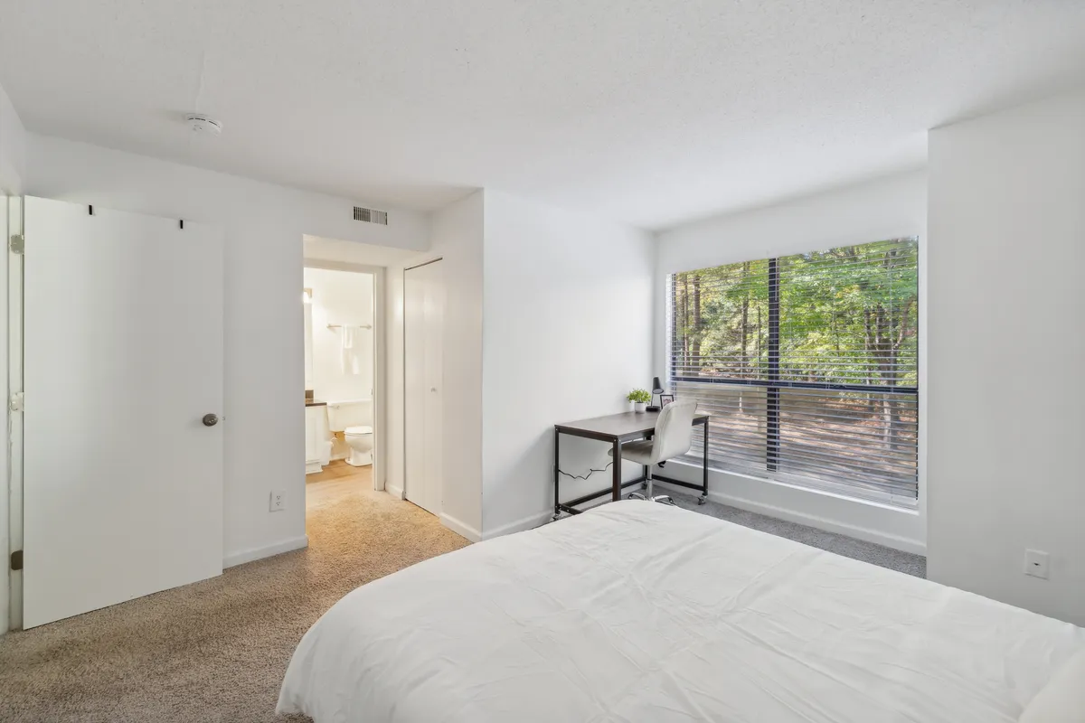 A spacious bedroom showcasing a cozy bed, a functional desk by the window, and peaceful views of nature outside. The room is designed for both relaxation and productivity, emphasizing versatility and comfort.