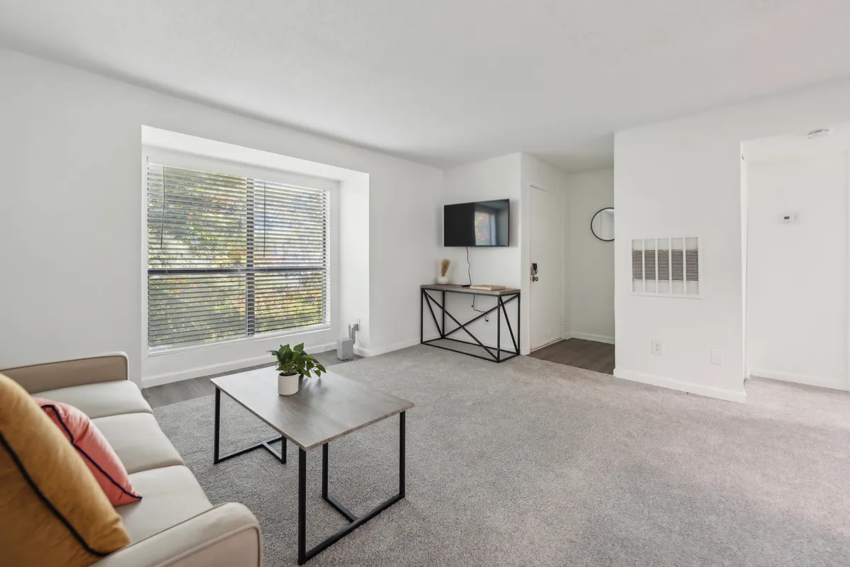 The image focuses on a calm living room with a sofa, coffee table, large window, and TV unit. Sunlight streams in, creating a bright, welcoming environment perfect for both relaxation and productivity.