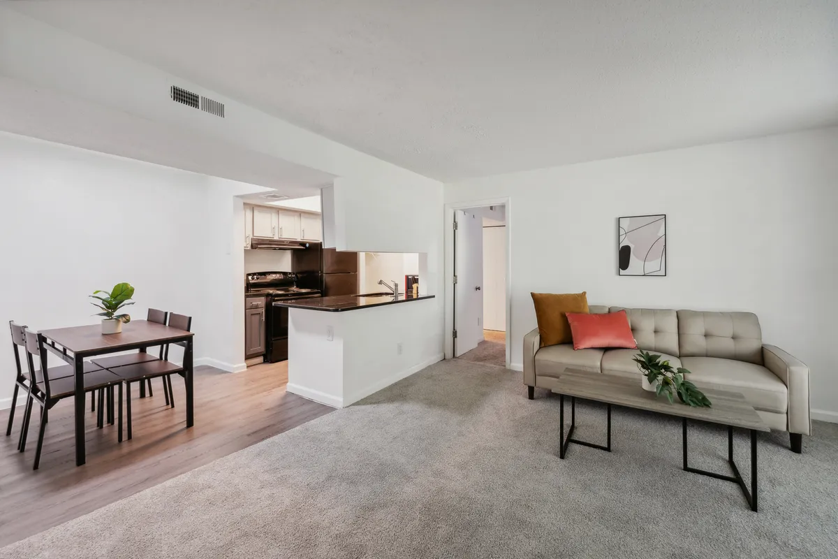This image captures a welcoming open-concept living area, featuring a cozy sofa with a cushion, a dining table ready for shared meals, and a view into the adjoining kitchen. The space is well-lit and styled to evoke comfort and relaxation, ideal for both unwinding after a long day and socializing with guests.