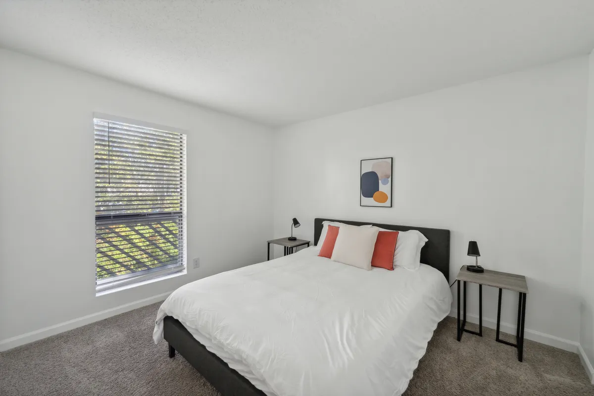 A tranquil bedroom with a comfortable bed positioned near a sunlit window, complemented by tasteful artwork and soft, inviting bedding. The space feels open and restful, perfect for unwinding or starting the day with natural light streaming in.