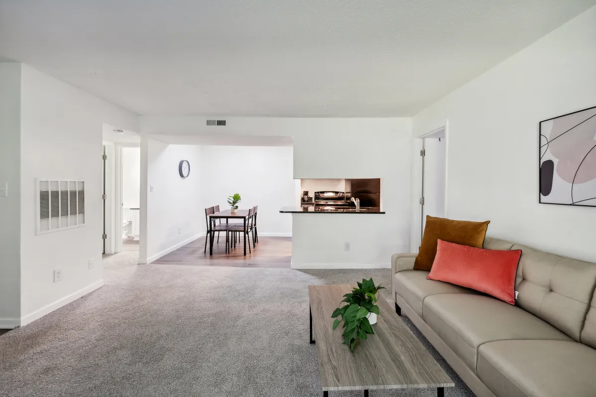 This image showcases a stylish, sunlit living area featuring a sofa with a bright orange pillow, a wooden coffee table adorned with a plant, and a dining area with chairs in the background. The arrangement suggests a comfortable space ideal for both relaxing at home and connecting with others.